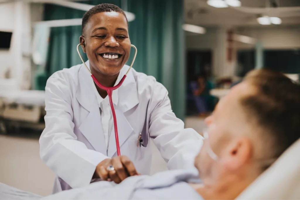 Smiling doctor using stethoscope on patient in hospital.